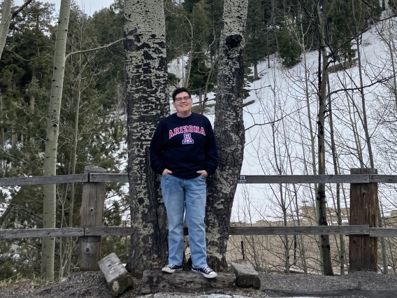 A picture of Francisco standing in front of trees in a winter landscape. They are smiling with their hands in their pockets, wearing a navy UA sweatshirt.