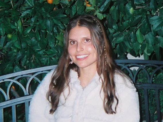 A photo of Emerson sitting on a bench in front of a wall of foliage in a fuzzy coat