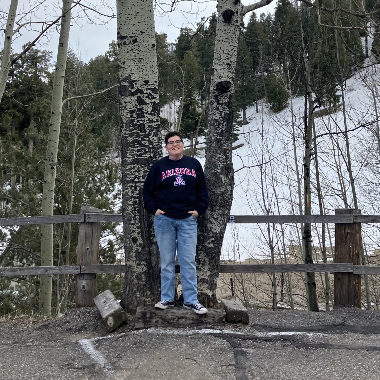 A picture of Francesca standing in front of trees in a winter landscape. They are smiling with their hands in their pockets, wearing a navy UA sweatshirt.