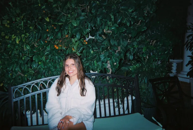 A photo of Emerson sitting on a bench in front of a wall of foliage in a fuzzy coat